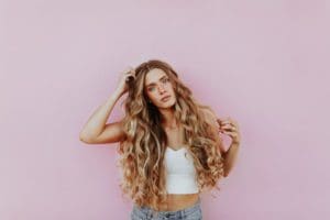 Woman with long curly hair wearing a crop tank and jeans in fron of pink background.